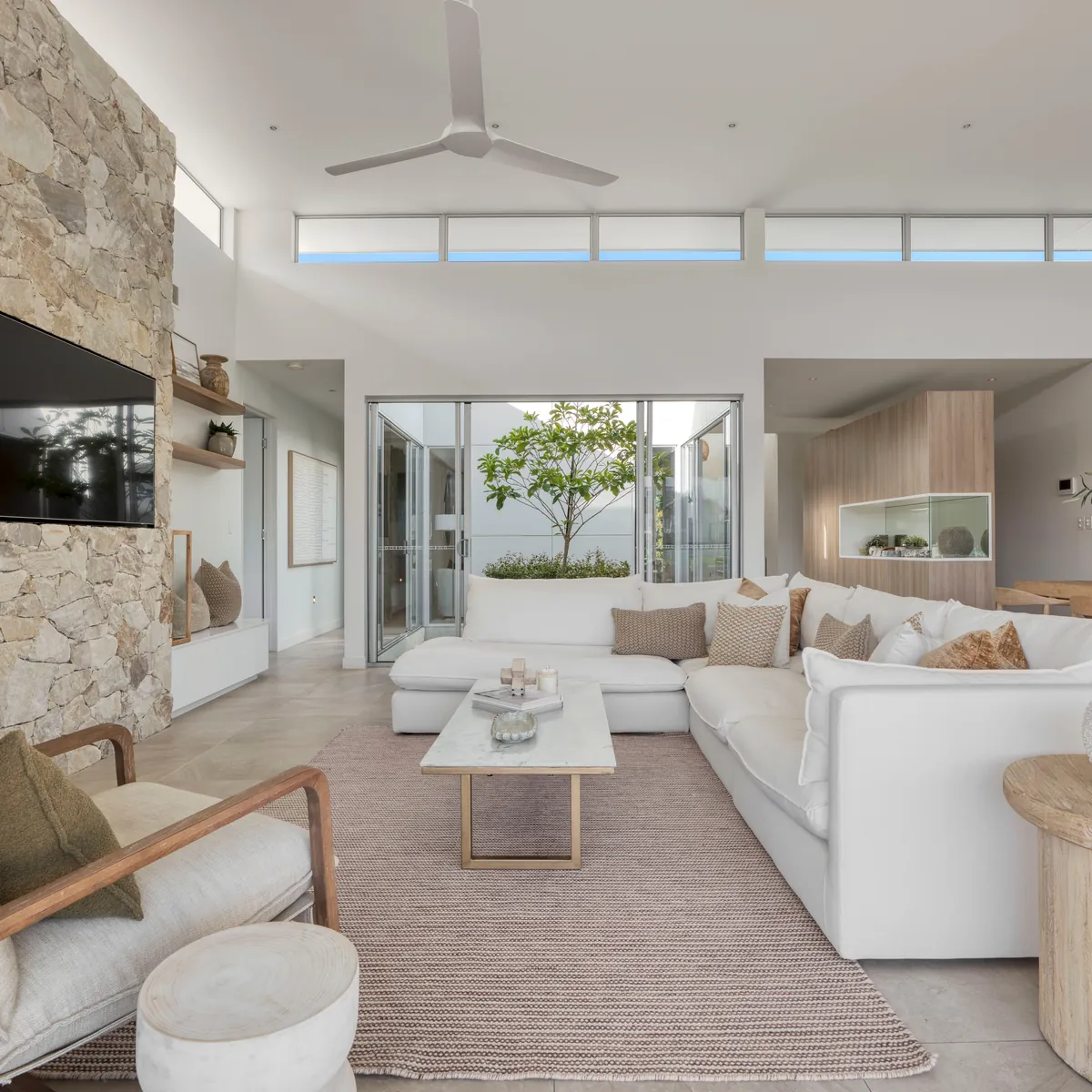 An open plan living room with a ceiling fan, recessed ceiling downlights, a lit display niche and a wall mounted television on a stone feature wall