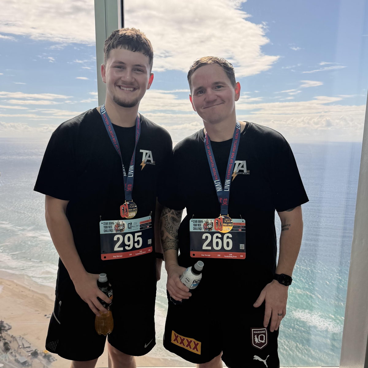 Tom and his apprentice Sam at the top of the Q1 tower in TA Electrical shirts, wearing their climb numbers and finisher medals, with the Gold Coast beach and ocean behind them